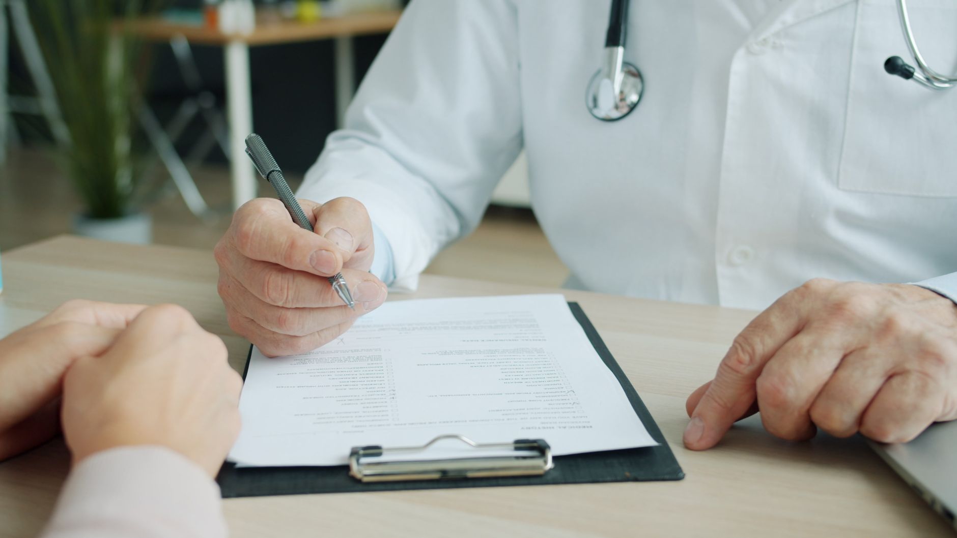 Médico preenchendo a ficha do paciente à mão, com a prancheta sobre a mesa entre os dois