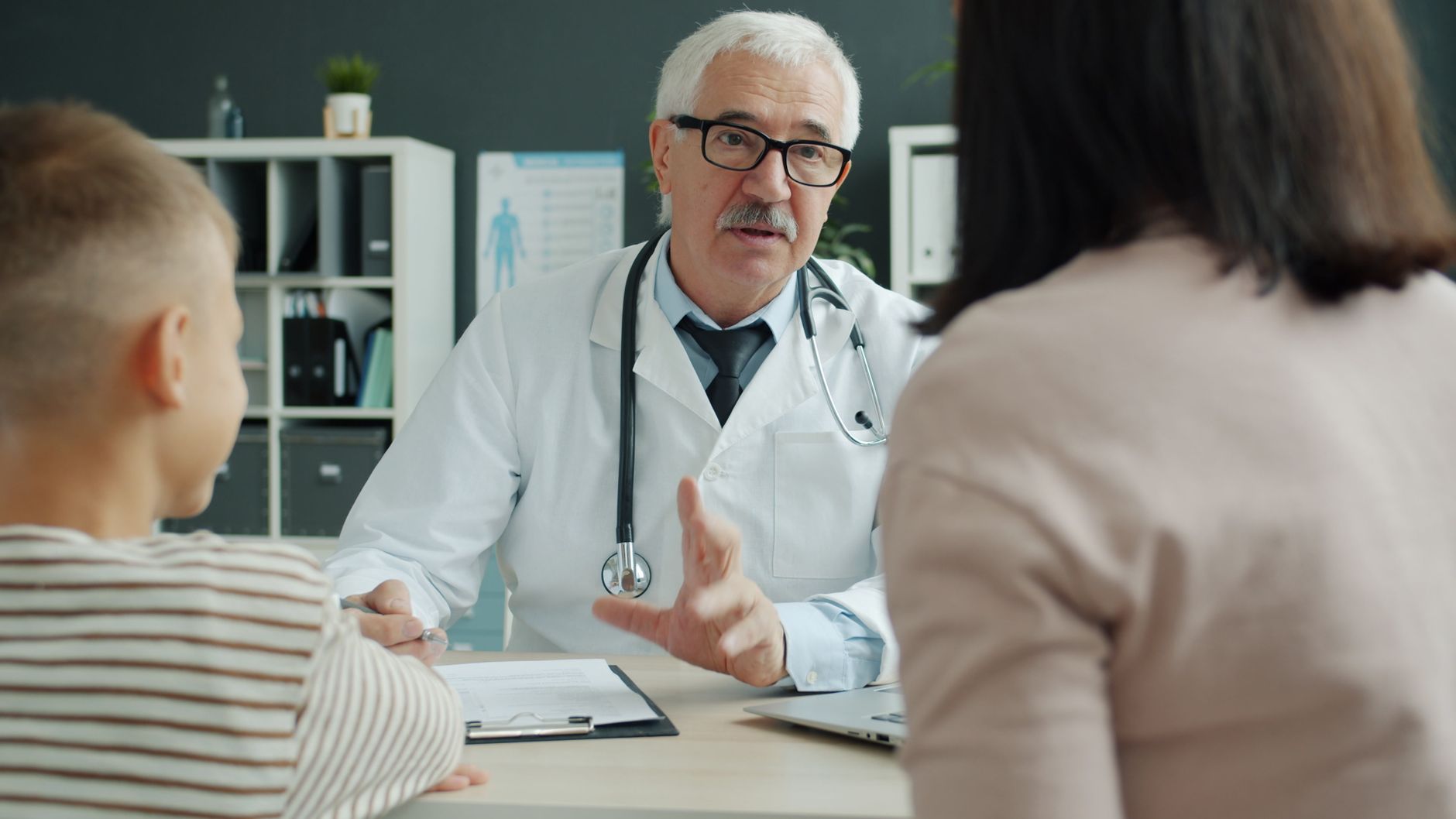 Médico conversando com uma mãe e um filho no consultório, com uma prancheta sobre a mesa